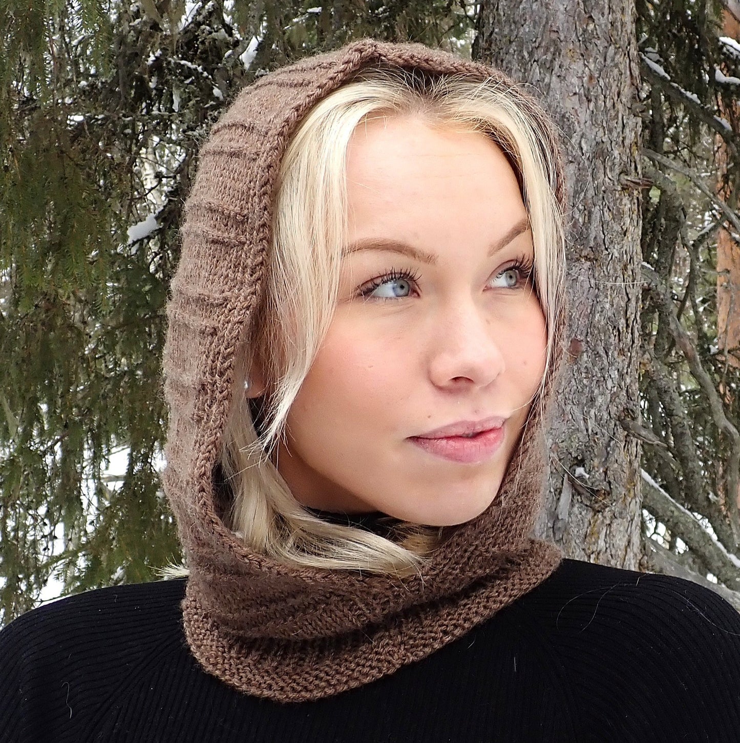 Vaaleahiuksinen nuori nainen seisoo vanhan kuusen edessä ja katsoo sivulle. Hänellä on yllään musta pusero ja ruskea neulottu huppukauluri. Huppukaulurin pinnassa on vaakasuoria kohoneuleraitoja.