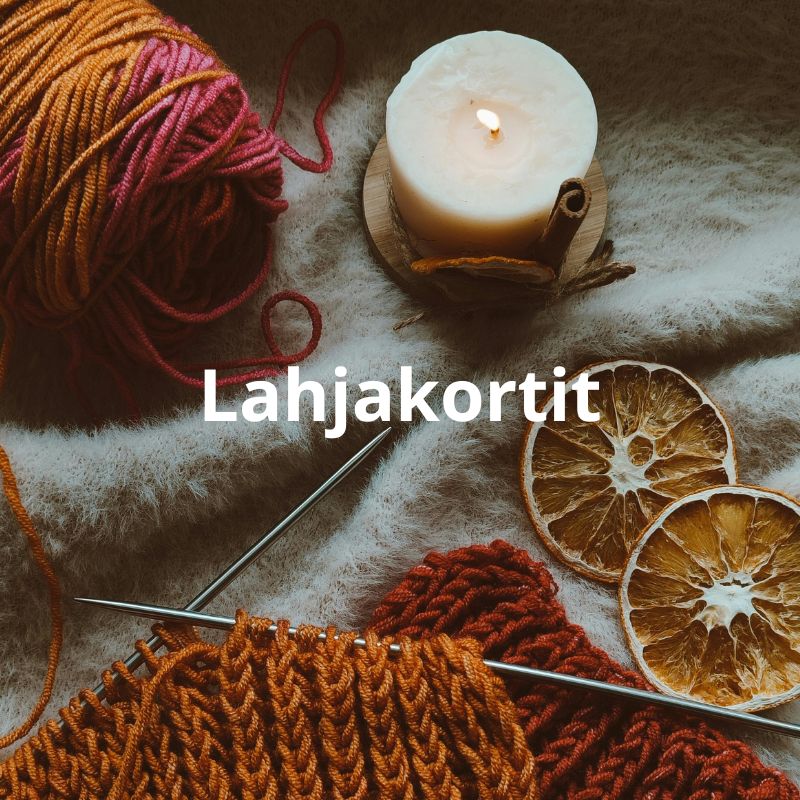 Vaalealla huovalla oranssisävyinen lankakerä, oranssi neule puikoilla ja punainen neule sen alla. Vieressä on kuivattuja appelsiininkuoria ja valkoinen palava kynttilä. Valkoisin kirjaimin teksti Lahjakortit.