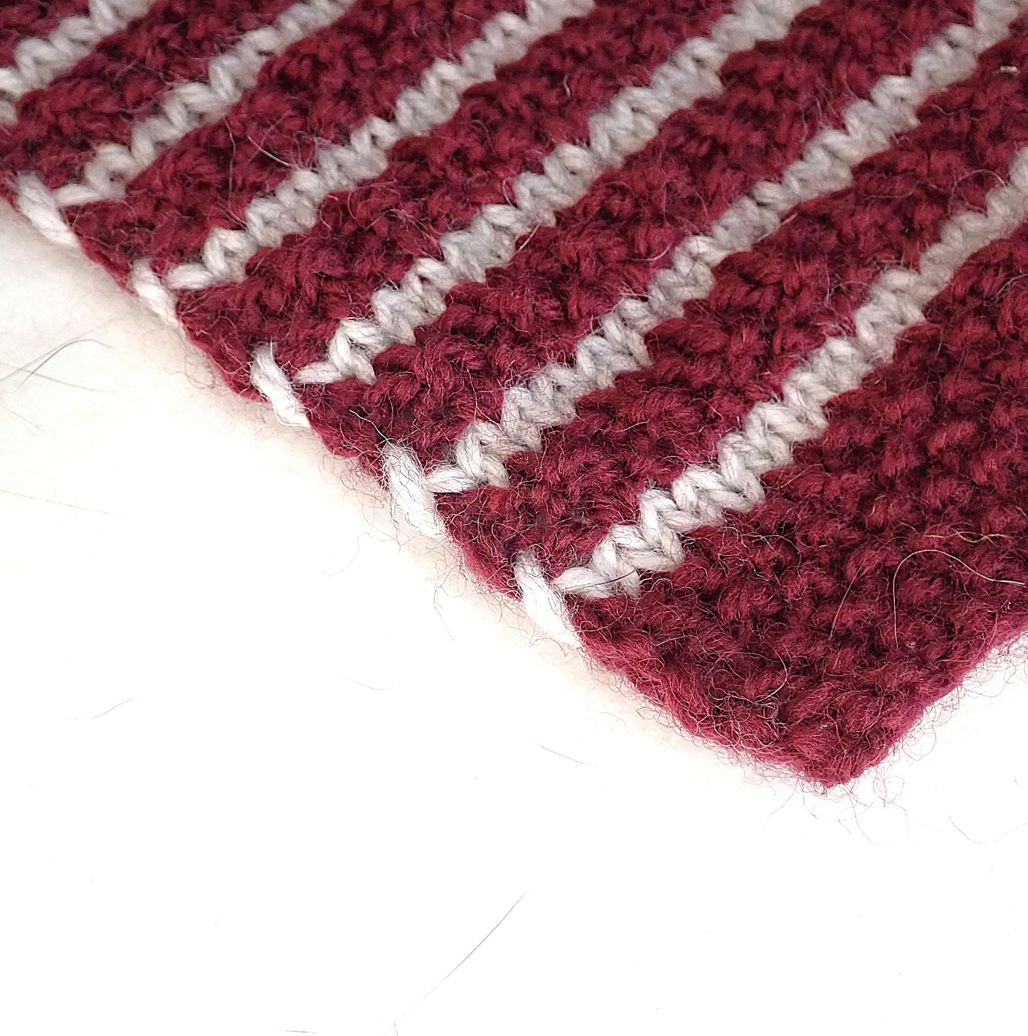 Neule, jossa on viininpunaisia helmineuleraitoja ja kapeita vaaleanharmaita raitoja. Neuleen reunassa eri väriset langat kiertyvät toistensa ympäri.