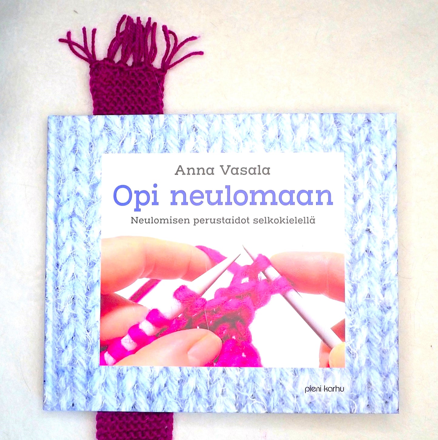 Viininpunainen minihuivi Opi neulomaan -kirjan välissä kirjanmerkkinä.