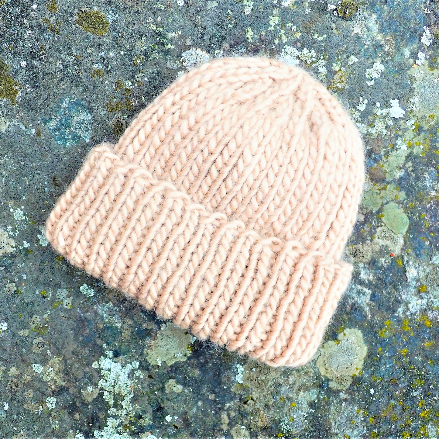 Harmaasävyisen kiven päällä on paksusta langasta neulottu muhkea beige pipo. Pipo on 1 oikein 1 nurin joustinneuletta.