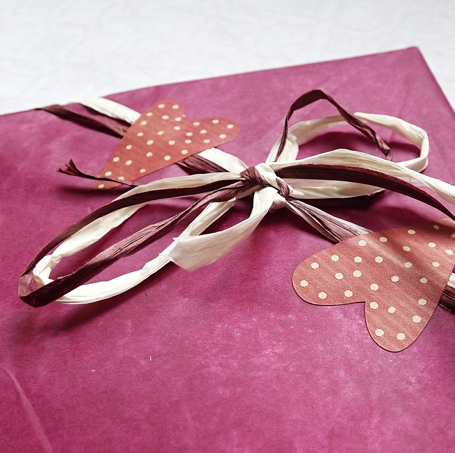 Selkosilmukat lahjapaketti. Viininpunaiseen silkkipaperiin kiedotun kirjan kulma. Paketti on sidottu tumman viininpunaisella ja beigellä paperinaurulla, joista on tehty rusetti. Lahjanarut on varmistettu viininpunaisilla sydäntarroilla, joissa on pieniä beigejä pisteitä.