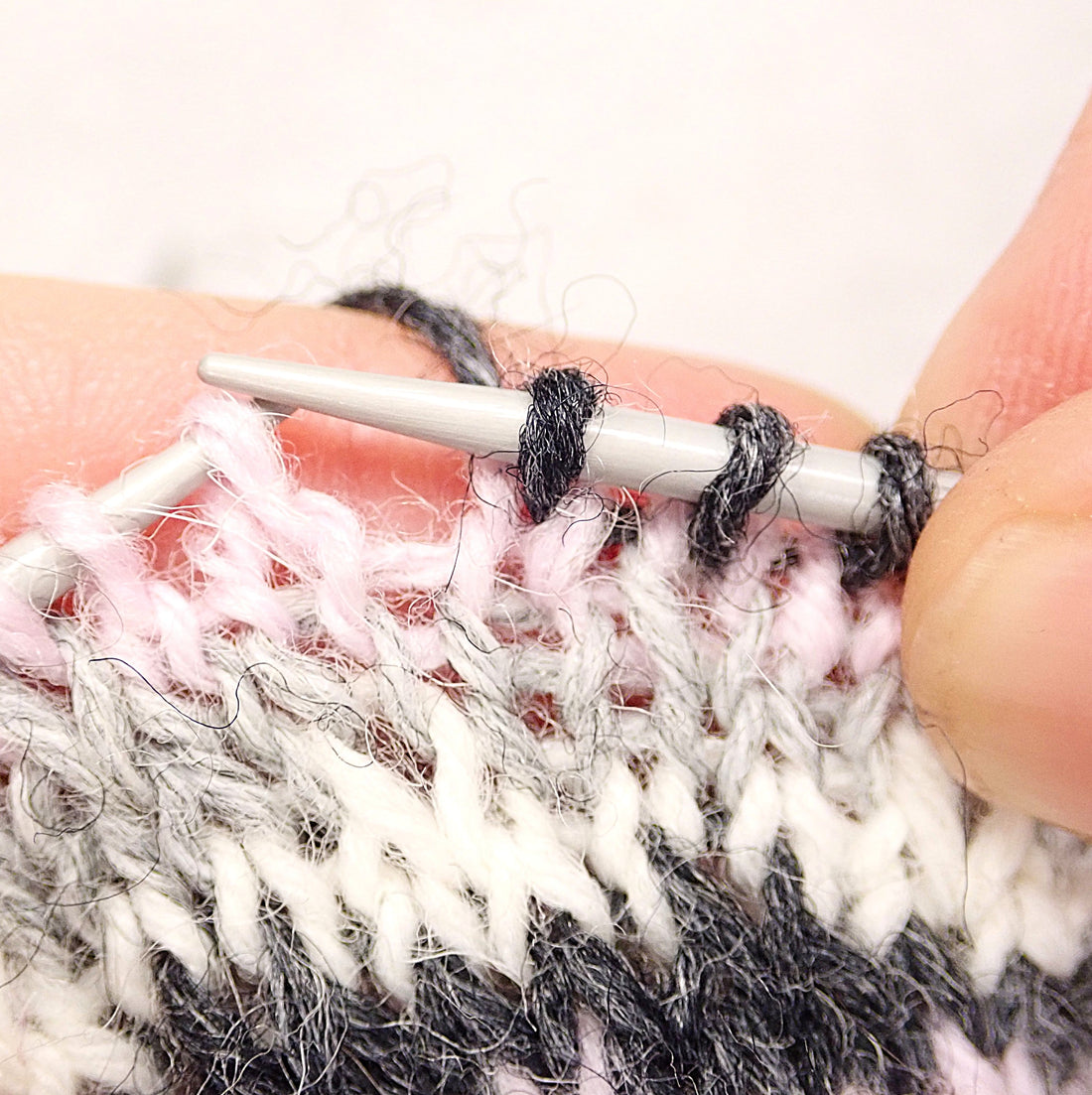 Kädet pitelevät puikkoja, joilla on keskeneräinen kirjoneule. Puikolle neulotaan juuri mustaa lankaa. Alempana on vaaleanpunaista, vaaleanharmaata ja luonnonvalkoista neuletta.