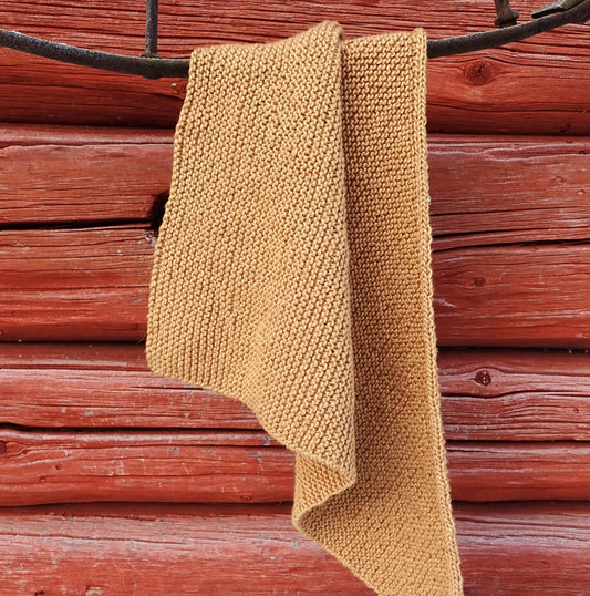 Kullankeltainen neulottu kolmiohuivi on laskostettu vanhan rautapyörän päälle. Taustalla punaista hirsiseinää.