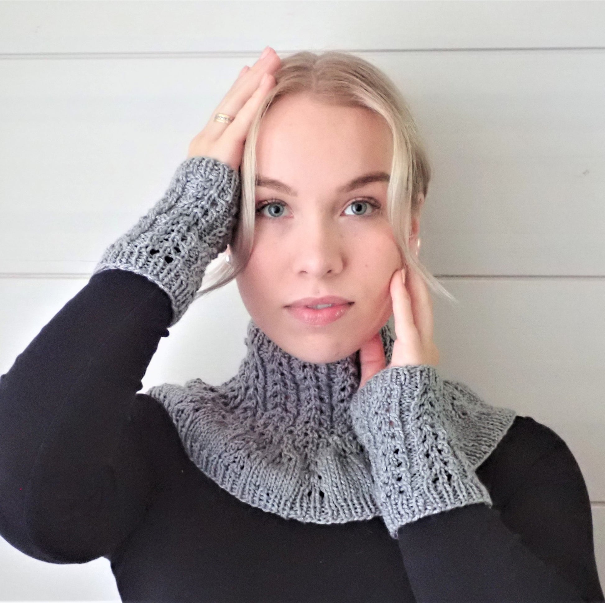 Nuori nainen, jolla  vaaleat pitkät hiukset ovat kiinni. Muutama hiussuortuva kehystää kasvoja. Naisella on yllään musta paita ja harmaa kaarrokekauluri pitsineuletta. Hän on nostanut kätensä lähelle kasvoja, jotta myös samanlaiset rannekkeet näkyvät hyvin.