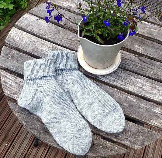 Harmaat neulotut villasukat kelottuneella puutarhapöydällä. Vieressä vanha alumiininen mittakannu, johon on istutettu sinisiä kukkia.