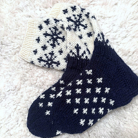 Sinivalkoiset kirjoneulevillasukat lampaantaljalla. Sukkien valkoisessa varressa on suuria tummansinisiä lumihiutalekuvioita. Sukkien tummansinisessä teräosassa on pieniä valkoisia lumihiutaleita.