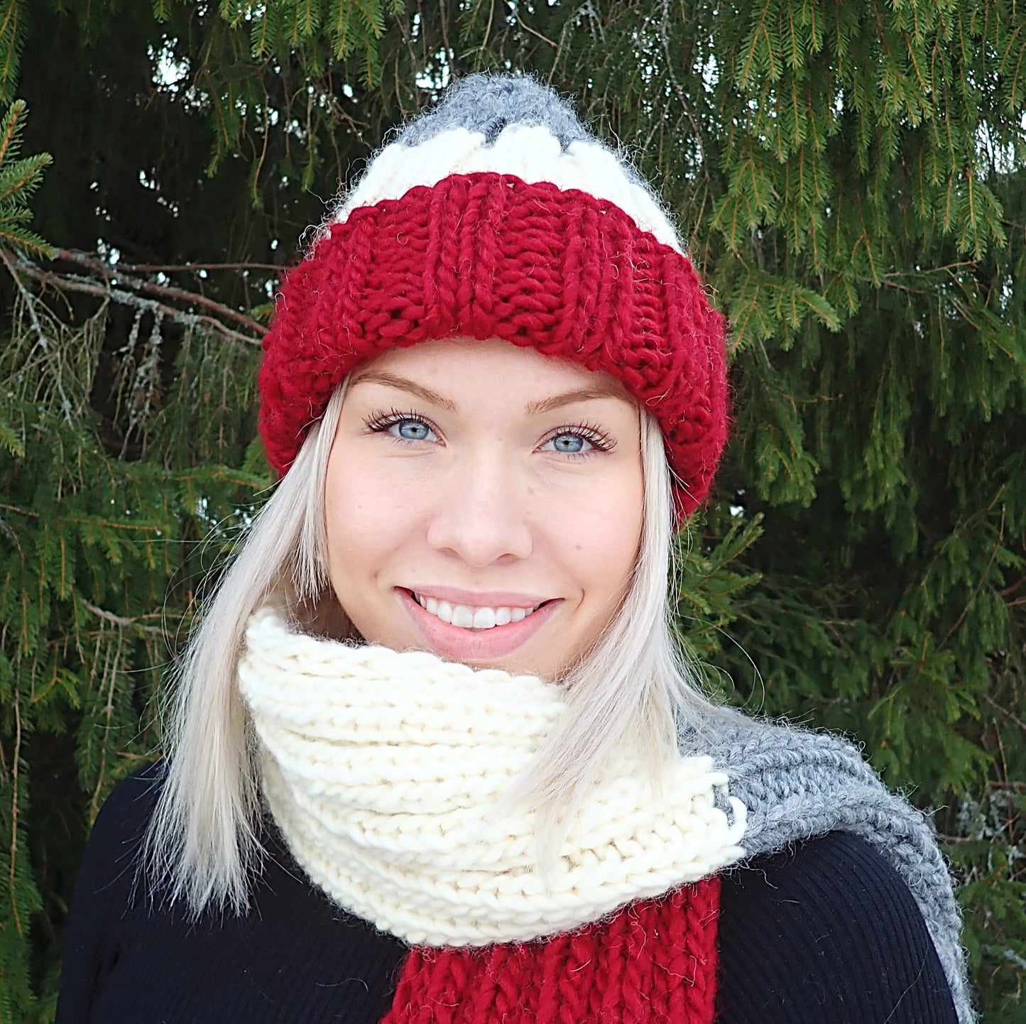 Vaaleahiuksinen nuori nainen hymyilee iloisesti. Hänellä on yllään muhkea pipo ja kaulahuivi, jotka ovat joustinneuletta. Neuleissa on leveät raidat tummanpunaista, luonnonvalkoista ja harmaata.