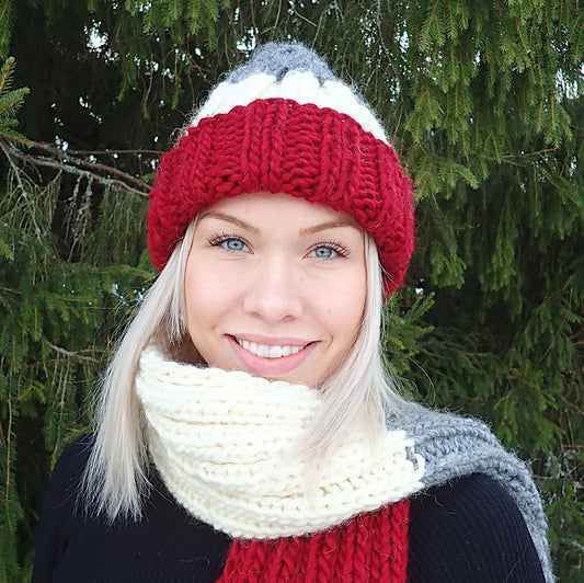 Vaaleahiuksinen nuori nainen hymyilee iloisesti. Hänellä on yllään muhkea pipo ja kaulahuivi, jotka ovat joustinneuletta. Neuleissa on leveät raidat tummanpunaista, luonnonvalkoista ja harmaata.