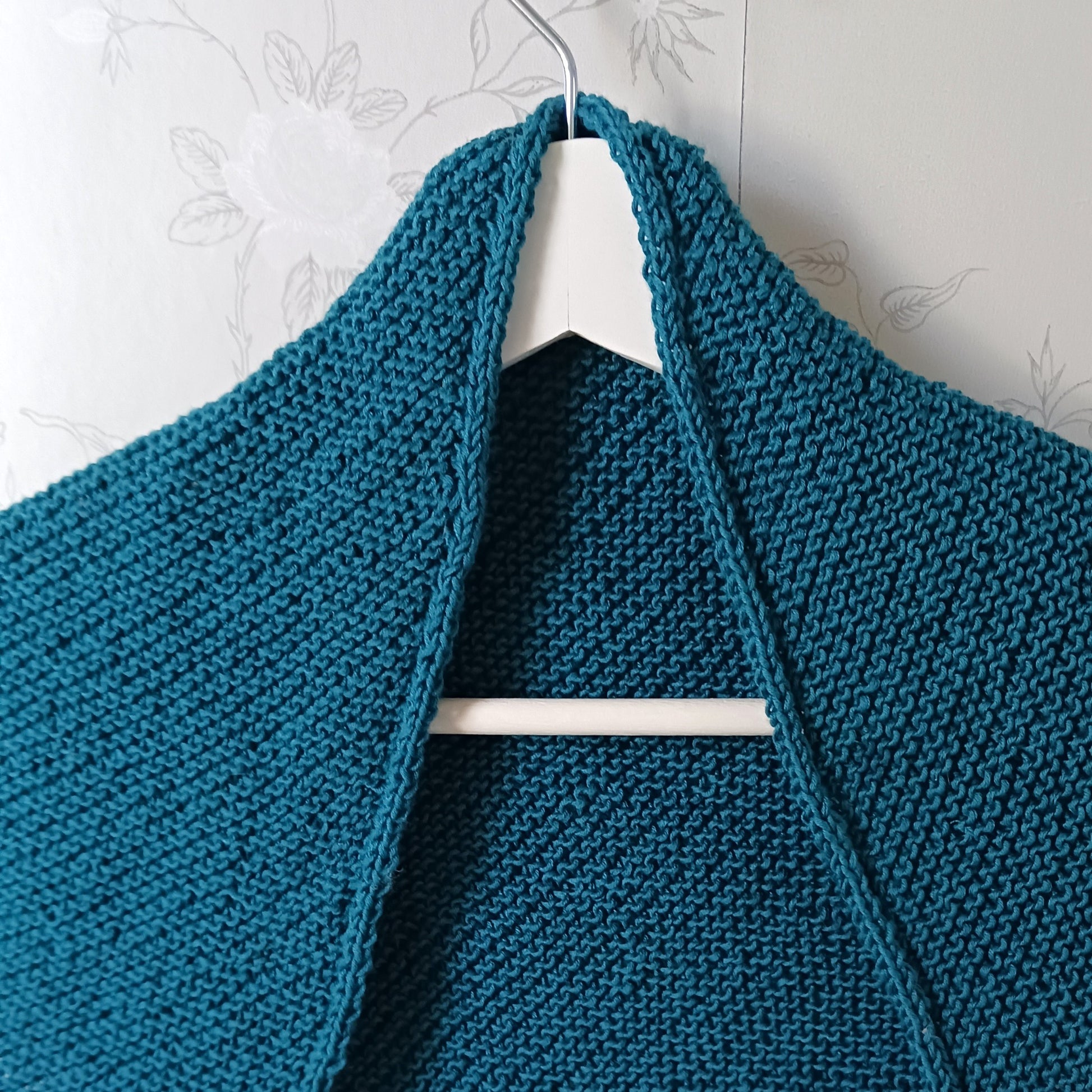 Petrolinsininen kolmiohuivi vaateripustimella. Huivi on ainaoikeinneuletta ja siinä om nyörireunus.