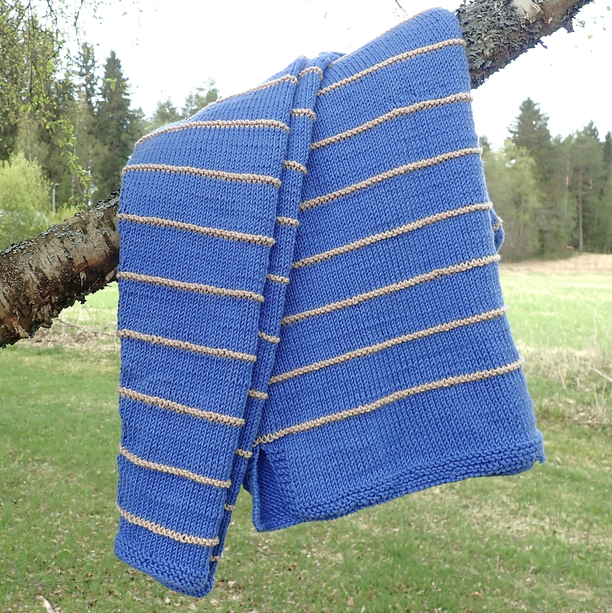 Taivaansininen neulepusero on laskostettu koivun oksalle. Puserossa on kapeita  beigejä kohoneuleraitoja. Taustalla näkyy vihreää nurmea, peltoa ja metsänreuna.