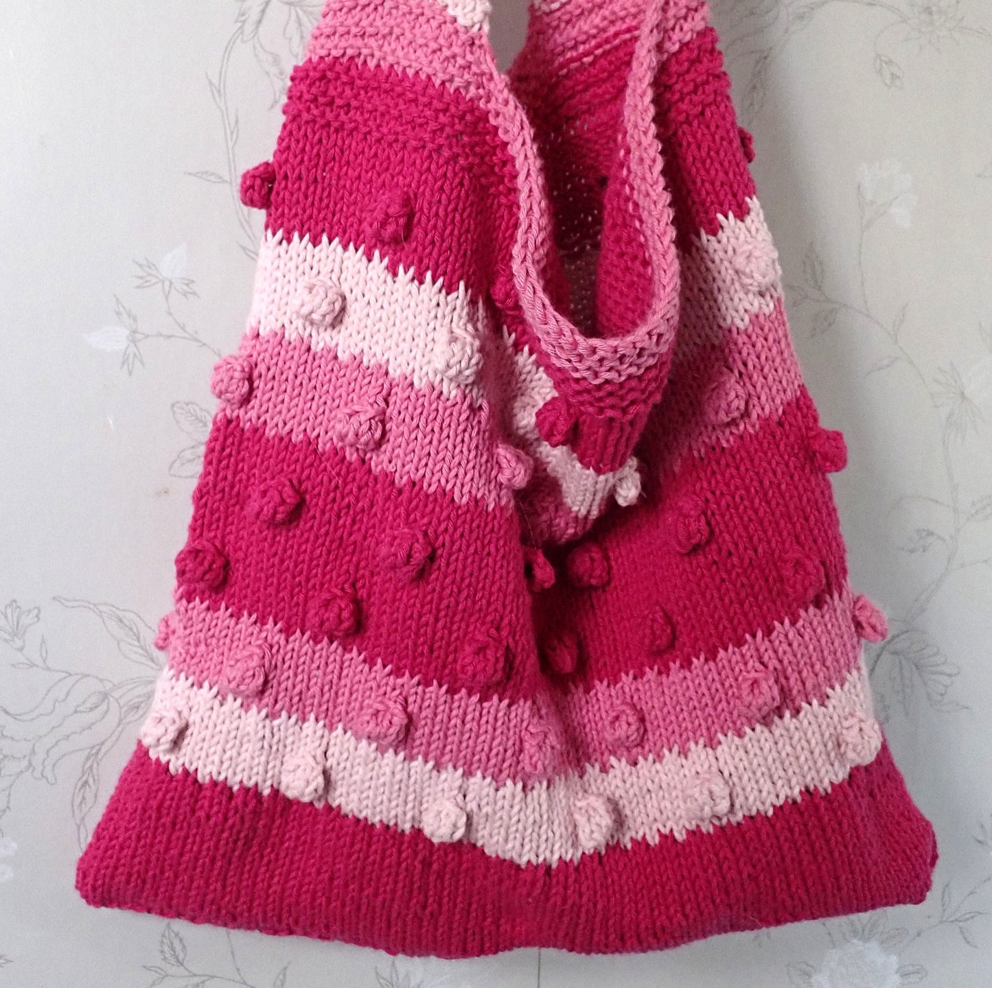 Valmis kesäkassi roikkuu naulakosta. Kassissa on raitoja, jotka ovat punaisen kolmea eri sävyä. Raitoihin on neulottu nyppyjä. Raitavärit vaihtuvat kuviolla, joka muistuttaa hammastusta. Kassin yläosa ja olkahihna ovat ainaoikeinneuletta.