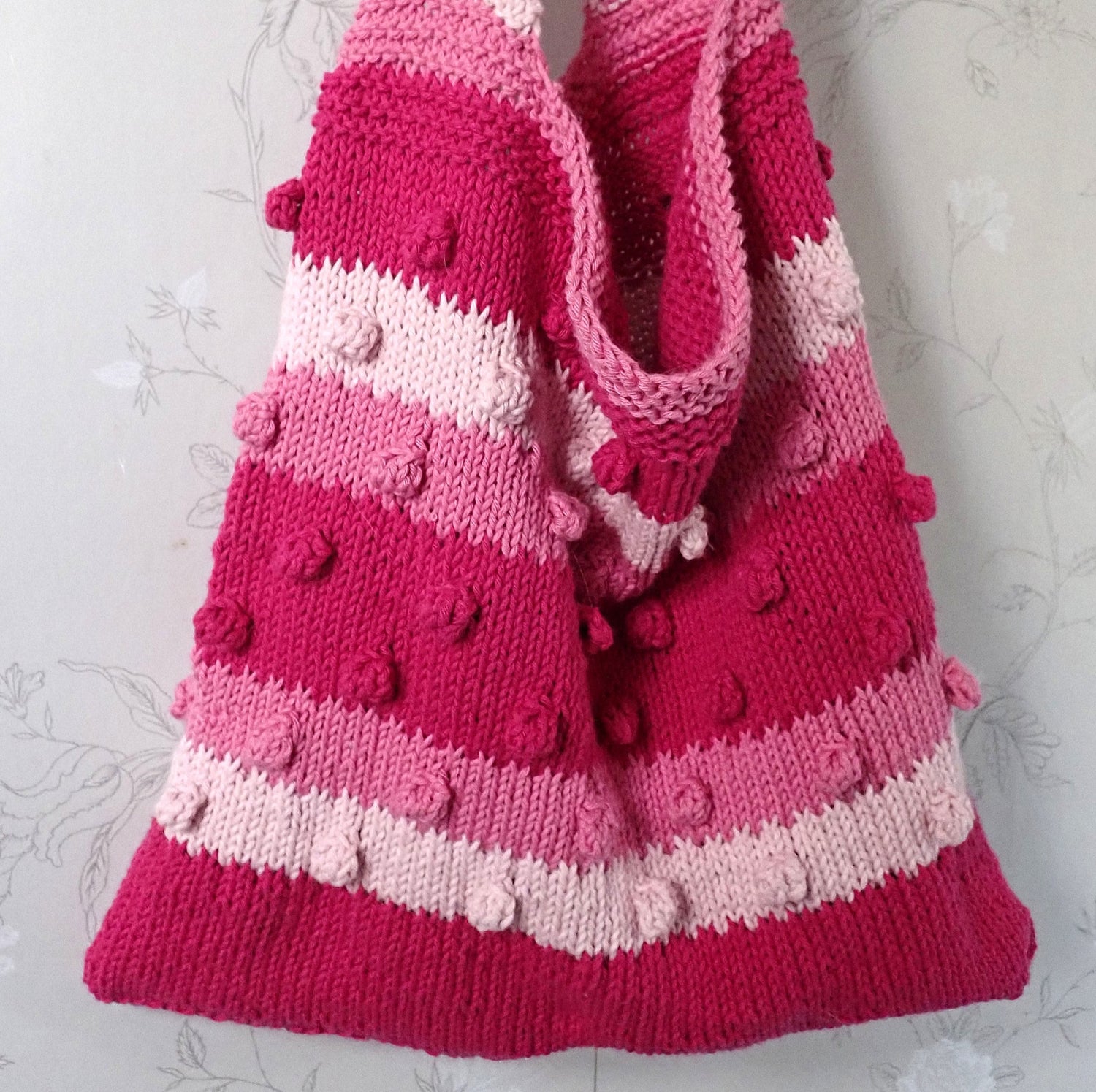 Valmis kesäkassi roikkuu naulakosta. Kassissa on raitoja, jotka ovat punaisen kolmea eri sävyä. Raitoihin on neulottu nyppyjä. Raitavärit vaihtuvat kuviolla, joka muistuttaa hammastusta. Kassin yläosa ja olkahihna ovat ainaoikeinneuletta.