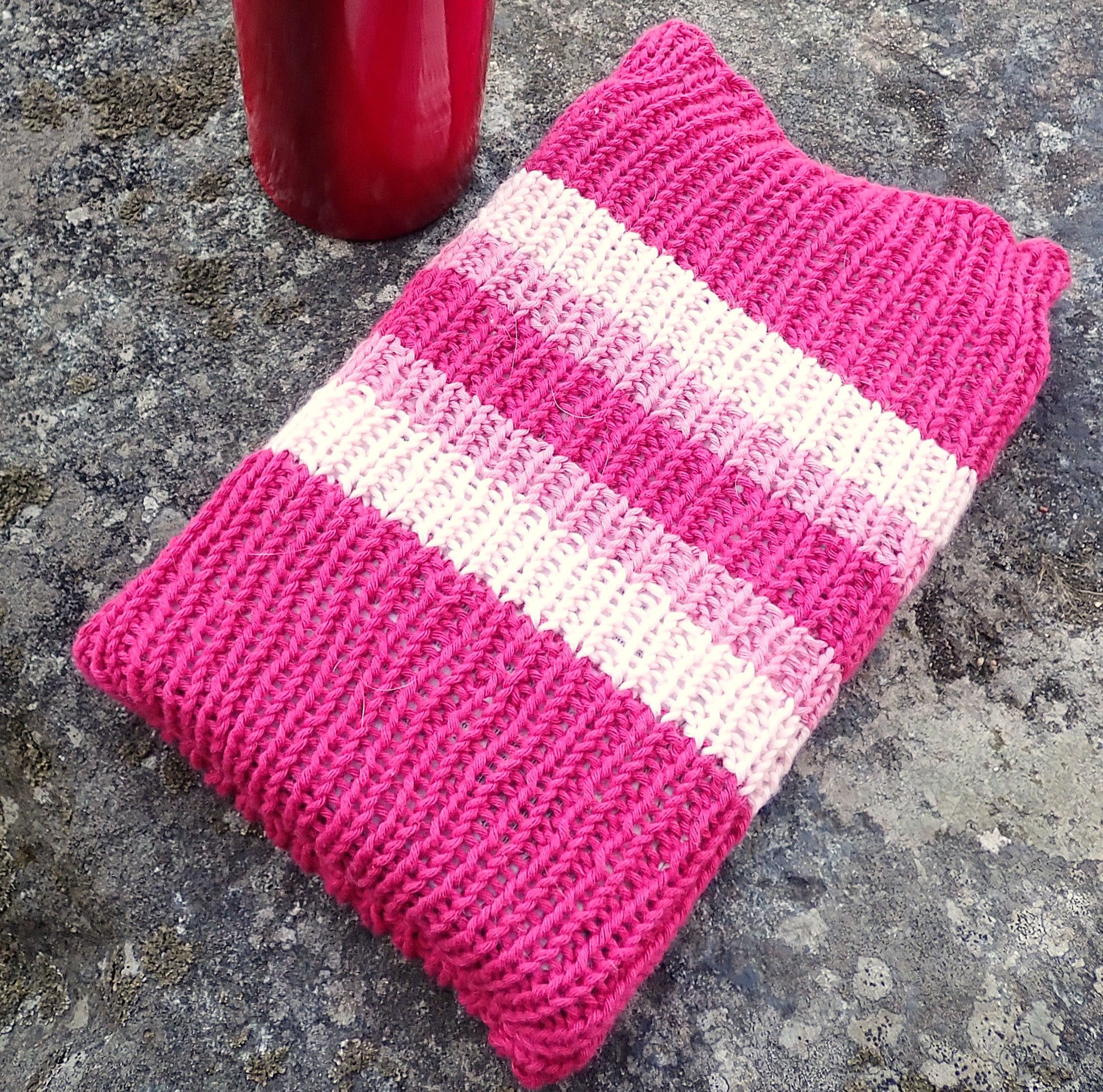 Suorakaiteen muotoinen joustinneulepussukka kiven päällä juomapullon vieressä. Pussukassa on fuksianpunaisia, vaaleanpunaisia ja vaalean pinkkejä raitoja.