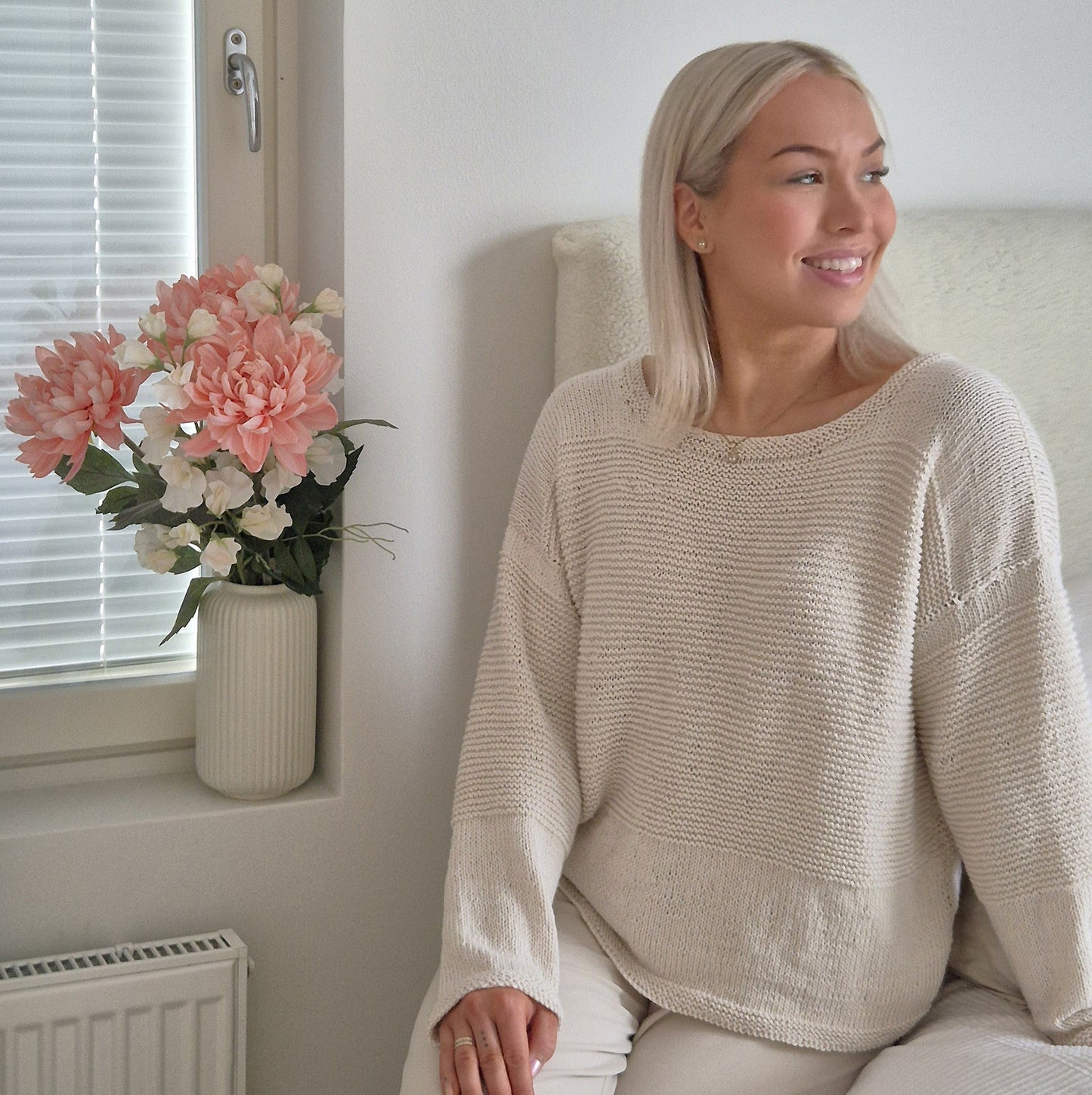 Nuori vaaleahiuksinen nainen, joka hymyilee. Hänellä on ylän luonnonvalkoinen neulepusero, jossa sileä neule ja ainaoikeinneule muodostavat pintoja. Puserossa on pyöreä pääntie. Ohje on tehty selkokielellä.