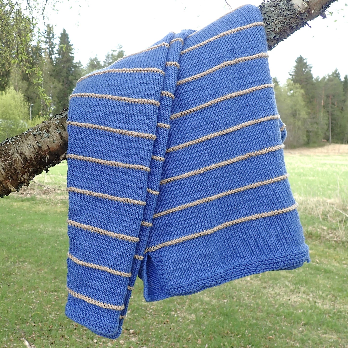 Taivaansininen neulepusero on laskostettu koivun oksalle. Puserossa on kapeita  beigejä kohoneuleraitoja. Taustalla näkyy vihreää nurmea, peltoa ja metsänreuna.