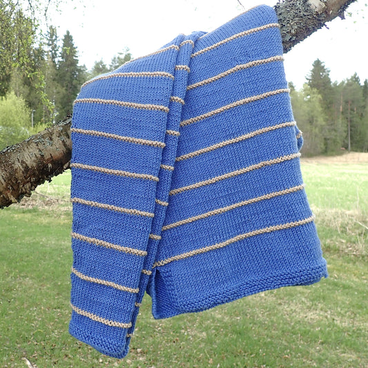 Taivaansininen neulepusero on laskostettu koivun oksalle. Puserossa on kapeita  beigejä kohoneuleraitoja. Taustalla näkyy vihreää nurmea, peltoa ja metsänreuna.