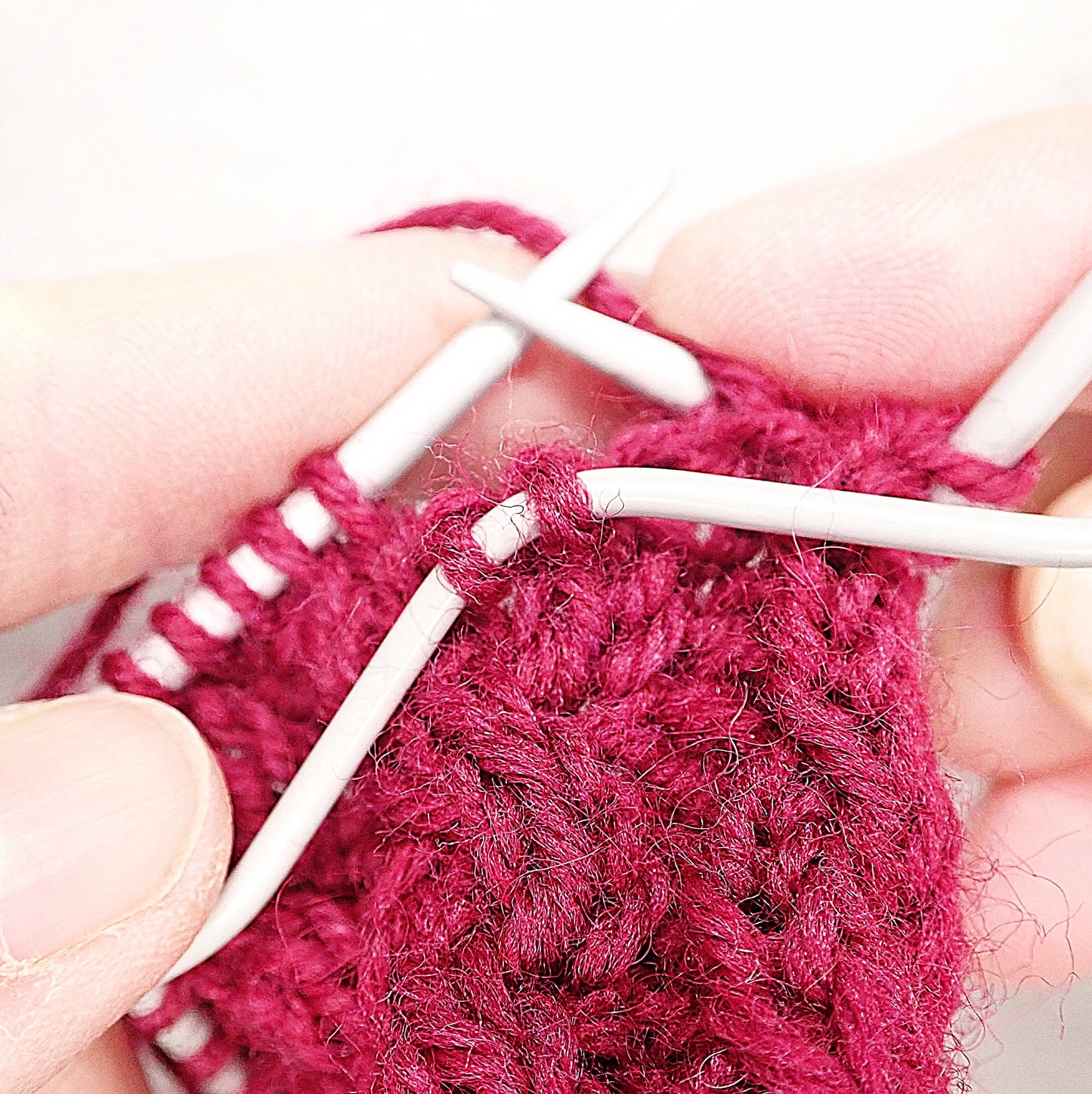 Puikoilla punainen neule, jossa tehdään palmikkoa. Kaksi silmukkaa on otettu palmikkopuikolle työn eteen.