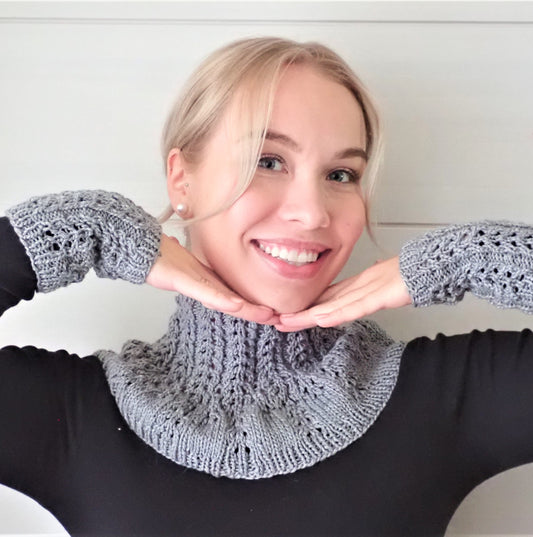 Nuori nainen, jolla  vaaleat pitkät hiukset ovat kiinni. Muutama hiussuortuva kehystää kasvoja. Naisella on yllään musta paita ja harmaa kaarrokekauluri pitsineuletta. Hän on nostanut kätensä lähelle kasvoja, jotta myös samanlaiset rannekkeet näkyvät hyvin.