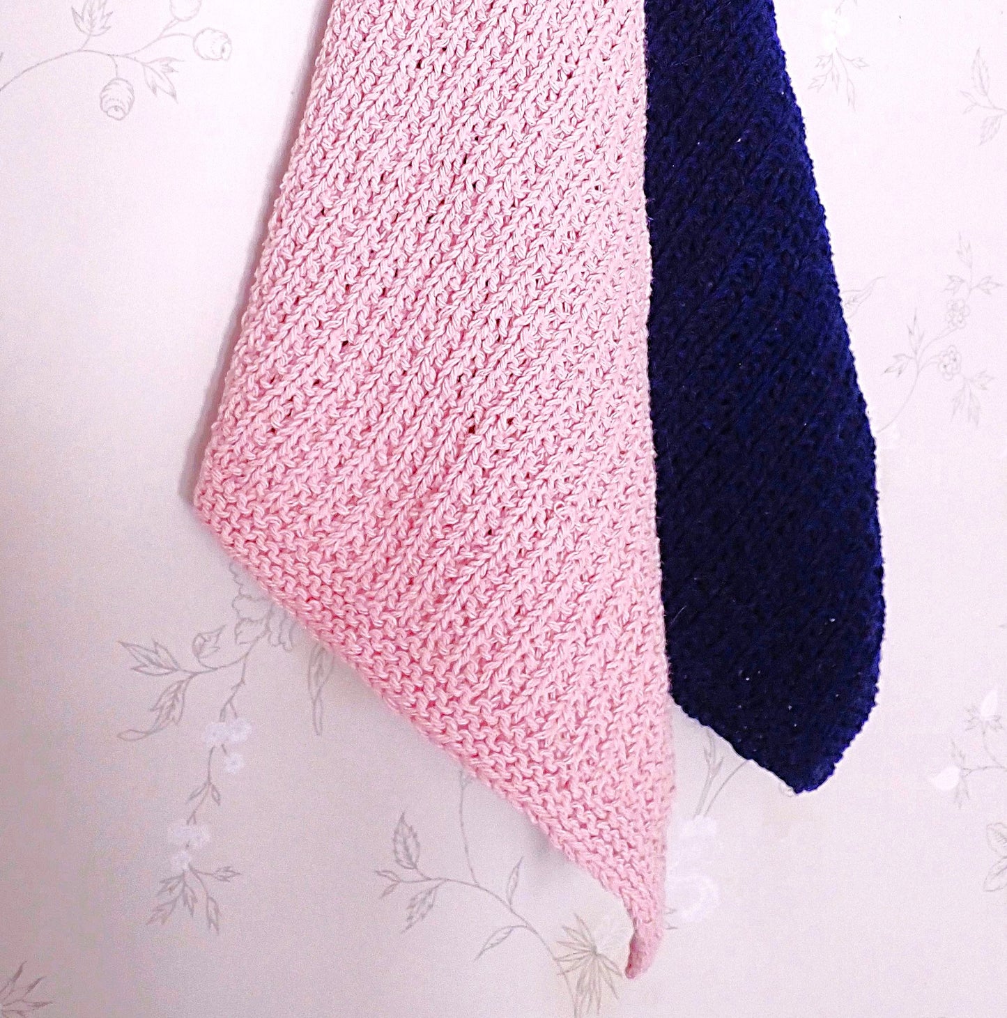 Seinällä roikkuu vaaleanpunainen ja tummansininen neulottu pyyhe. Pyyhkeiden pinta on elävän näköinen. Seinällä on harmaa tapetti, jossa on hentoja valkoisia kukkakuvioita.