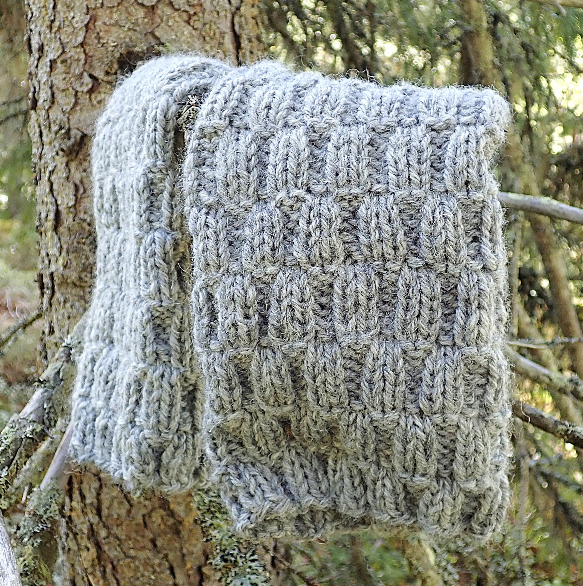 Harmaat säärystimet on taiteltu kuusen oksalle. Säärystimien pintaneule muistuttaa punotun korin kuviota. Taustalla havumetsässä paistaa aurinko.