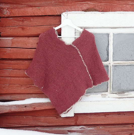Viininpunainen poncho vaateripustimessa vanhan, punaisen hirsirakennuksen ruutuikkunan edessä. 