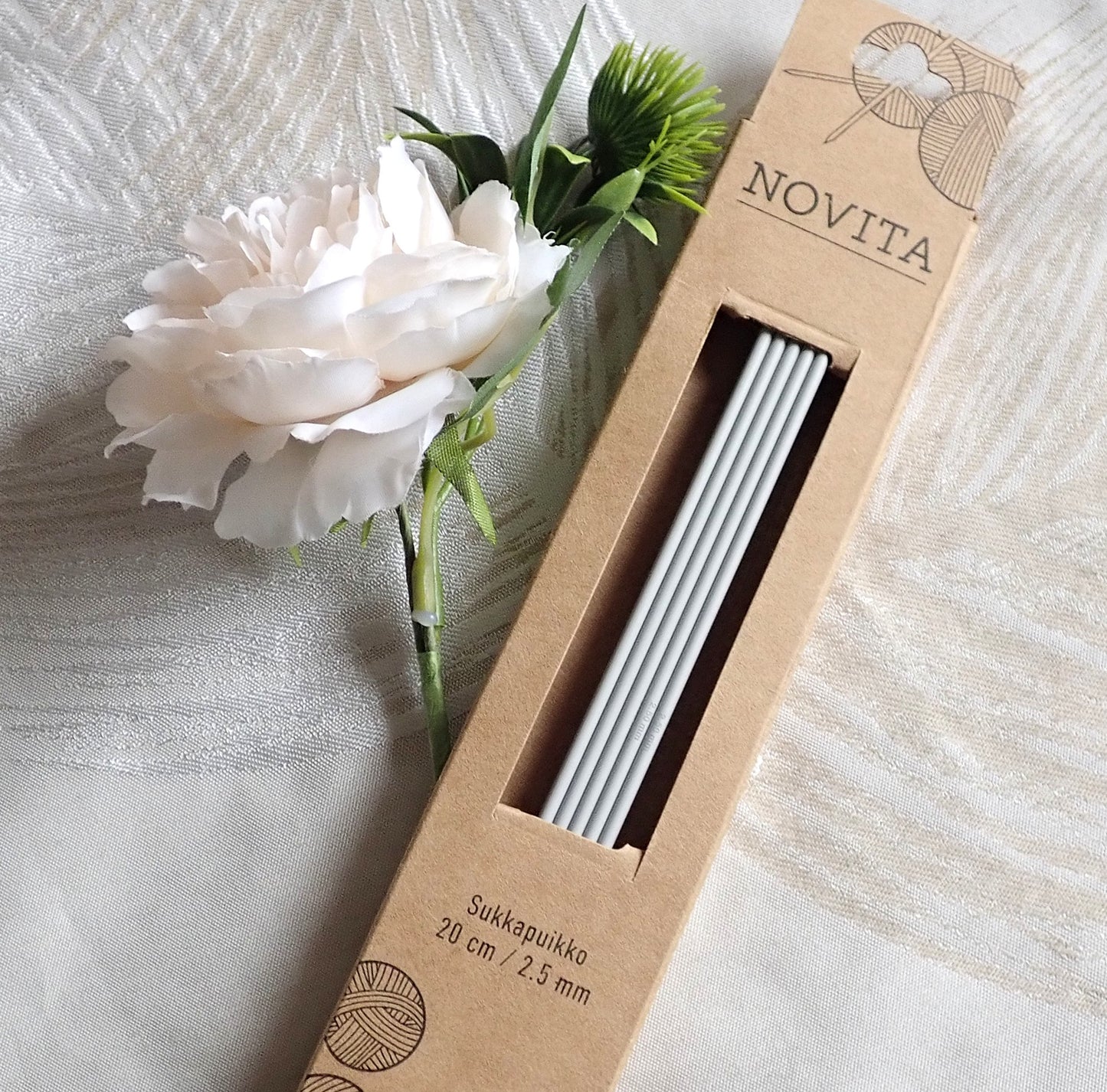 Novitan sukkapuikot kokoa 2,5 kartonkipakkauksessa. Vierellä valkoinen pioninkukka.