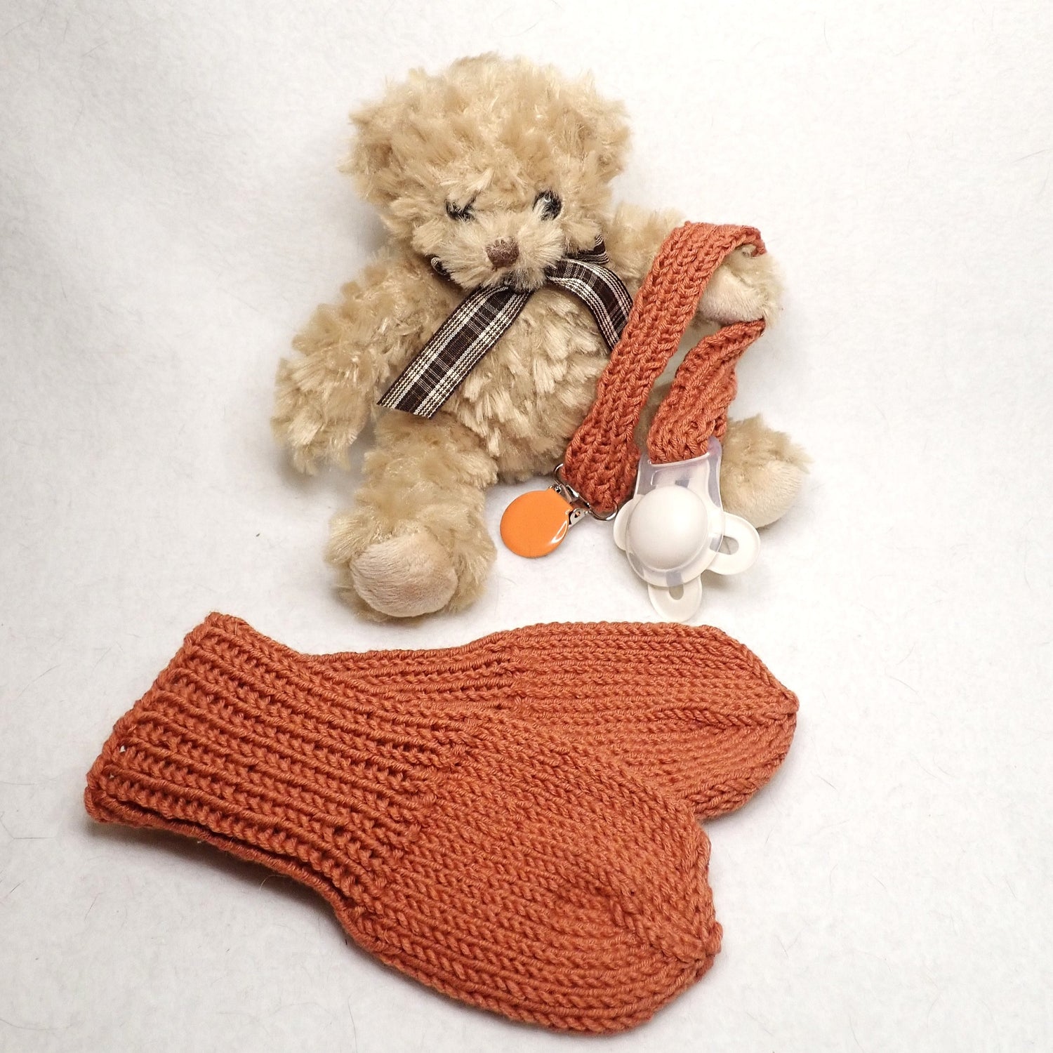 Oranssit vauvan neulotut lapaset, joissa ei ole peukaloa. Nalle pitelee neulottua tuttinauhaa, jossa oranssi klipsi.
