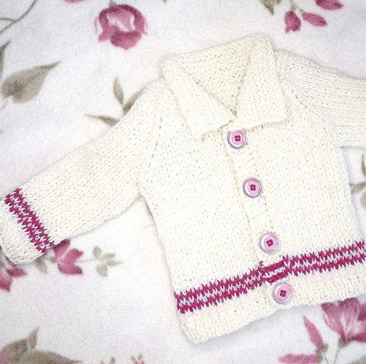 Luonnonvalkoinen vauvan villatakki lepää kukkakuvioisella viltillä. Takin helmassa ja hihansuissa on vaalean- ja fuksianpunaiset raidat. Takin pinta on patenttineuleen tapaista kohoneuletta ja takissa on leveä kaulus sekä vaaleanpunaiset napit.