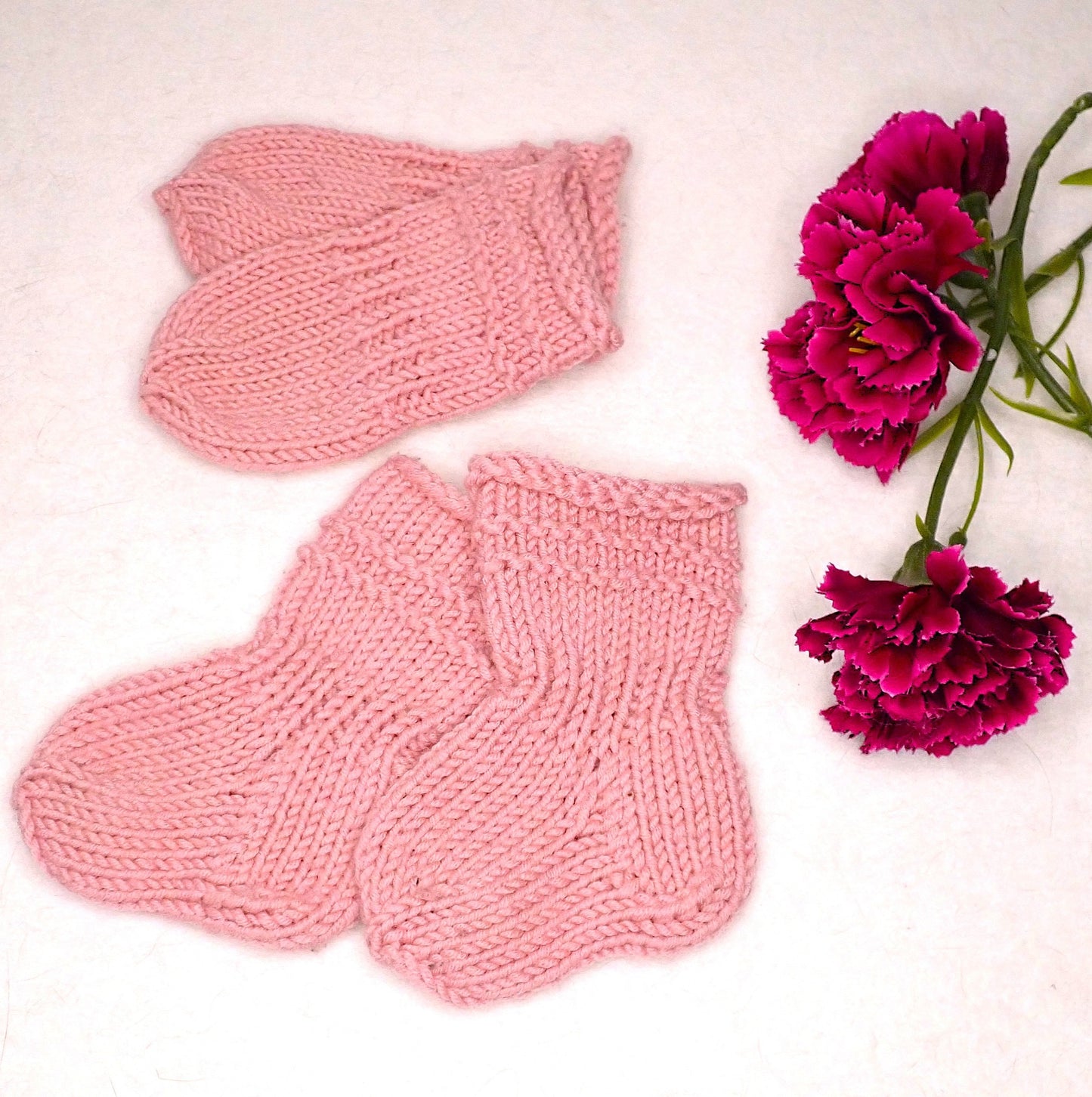 Vaaleanpunaiset vauvan lapaset ja sukat, joiden vieressä on kolme viininpunaista neilikkaa. Asusteiden varressa on tehosteena 2 kohoneuleraitaa.