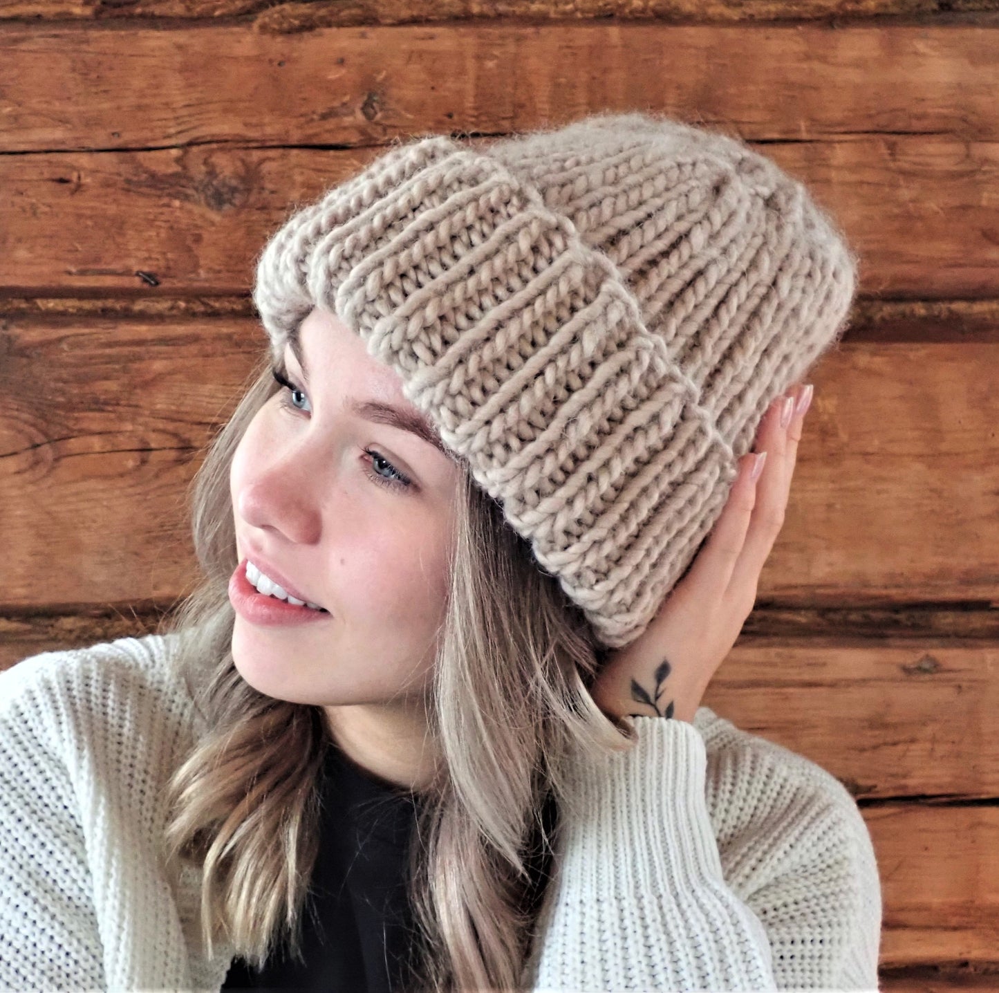 Nuori nainen hymyilee ja koskettaa kevyesti kädellään muhkeaa beigeä joustinneulepipoa, joka hänellä on päässään. Taustalla tummaa hirsiseinää.